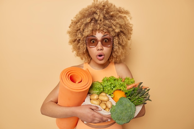 Shocked curly haired young woman stares impressed cannot believe own eyes carries fresh vegetables and rolled karemat leads healthy lifestyle wears sunglasses isolated over brown background