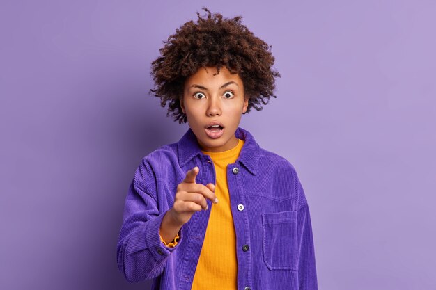 Shocked curly haired young woman stares with stunned expression indicates index finger directly at you holds breath from wonder dressed in fashionable outfit.