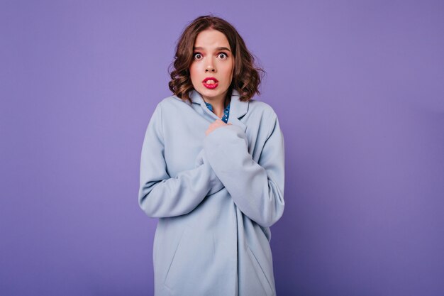 Shocked curly woman posing in coat in cold day. Indoor photo of emotional caucasian female model with wavy hairstyle isolated on purple wall.