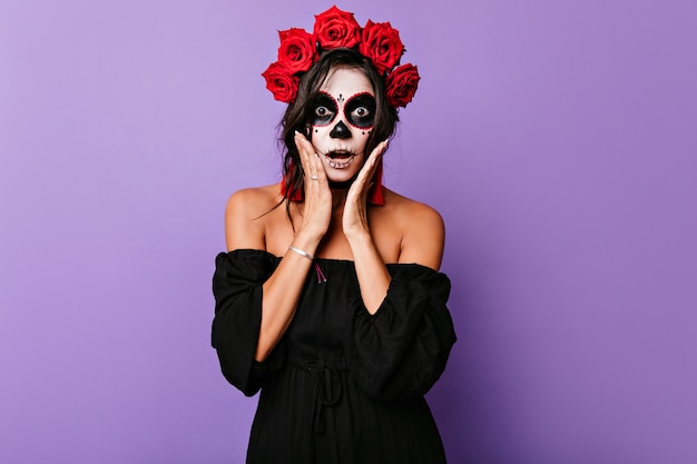 Shocked dark-eyed lady posing with mouth open in halloween. indoor shot of frightened female zombie with roses in hair.