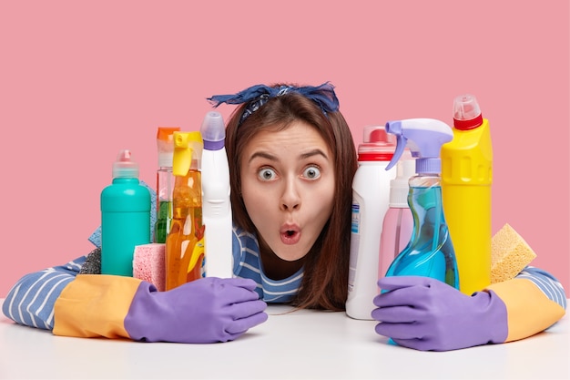 Free photo shocked dark haired lady with widely opened eyes, terrified facial expression, hugs bottles of solvent and spray, stupefied with much housework