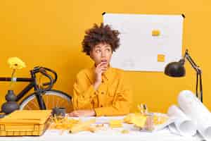 Free photo shocked dark skinned afro american female student works on blueprints dressed in yellow jacket analyzes drawbacks and corrects mistakes in drawings analyzes construction plan looks surprised aside
