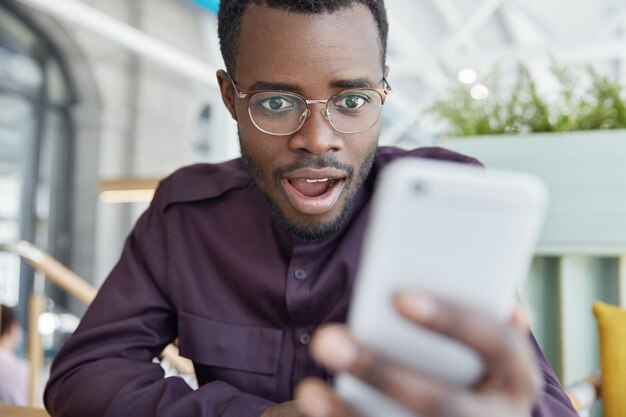 Shocked dark skinned businessman in eyewear, recieves notification on modern smart phone, gets notice to pay bills, has surprised expression.