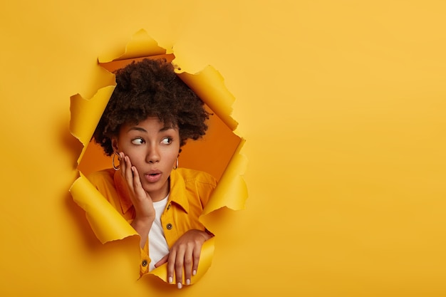 Free photo shocked dark skinned female model looks aside with embarrassement, holds palm on cheekbone, surprised by big discounts, wears fashionable clothes, poses in paper hole, yellow studio background