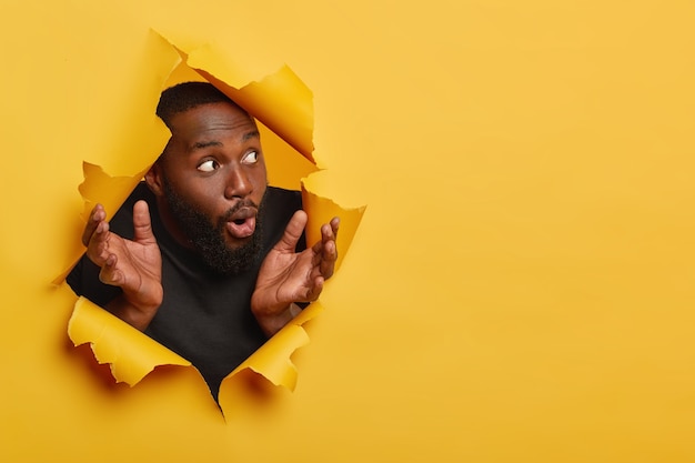 Shocked dark skinned man spreads palms sideways, reacts with surprisement, looks aside, wears black clothes, focused aside with impressed expression, poses in torn paper hole, yellow background