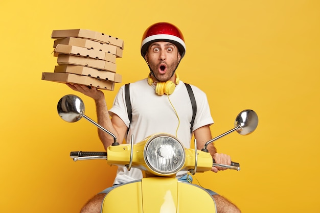 Shocked deliveryman with helmet driving yellow scooter while holding pizza boxes