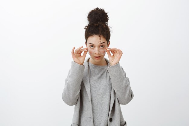 shocked disbelieving attractive european girl with curly hair combed in bun, taking off glasses, looking from under forehead focused and intrigued