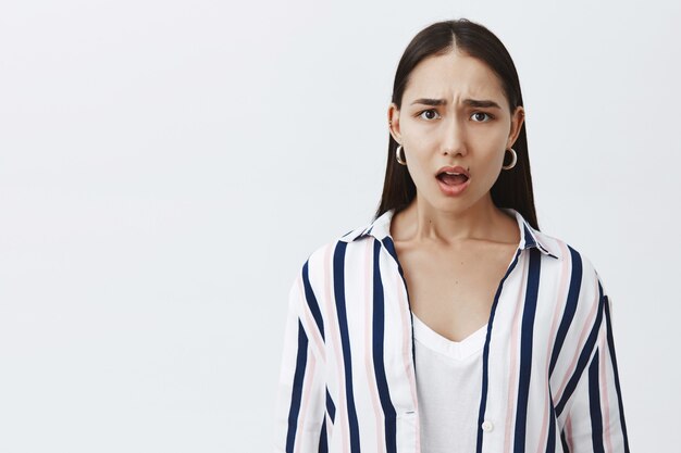 shocked displeased attractive young woman in striped blouse with natural beauty, frowning and gasping from disappointment, expressing empathy to poor friend, posing over gray wall