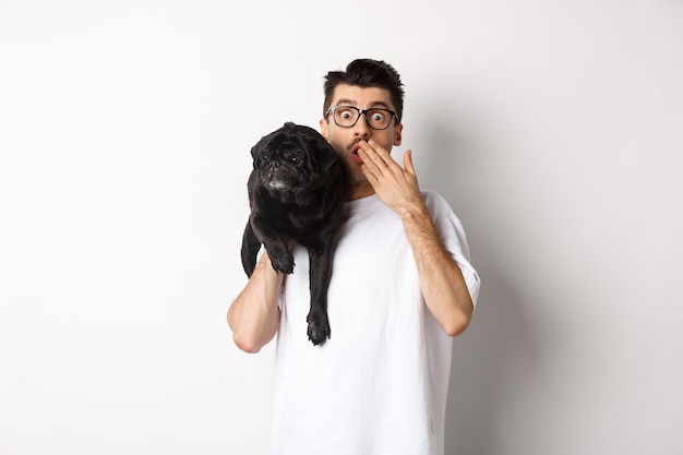 Shocked dog owner staring at camera, holding puppy on shoulder and gasping amazed. Handsome young man carry pug and reacting to promo advertisement, white background.