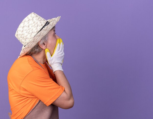 Shocked elderly female gardener wearing gardening hat and gloves puts hands on mouth and looks at side on purple