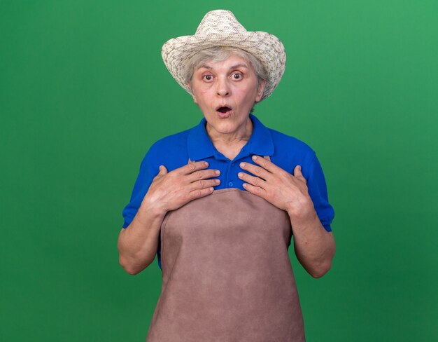 Shocked elderly female gardener wearing gardening hat puts hands on chest