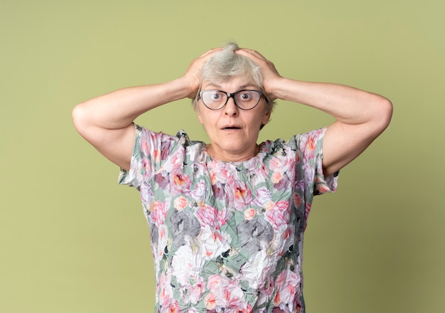 Free photo shocked elderly woman in optical glasses holds head isolated on olive green wall