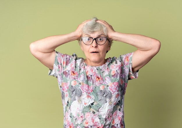Shocked elderly woman in optical glasses holds head isolated on olive green wall