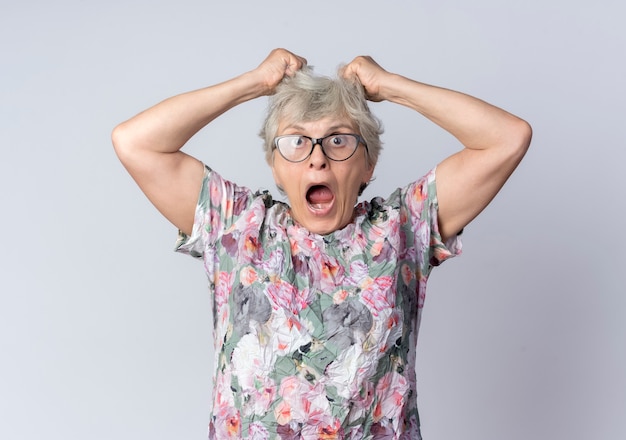 Free photo shocked elderly woman in optical glasses holds and lifts hair looking isolated on white wall