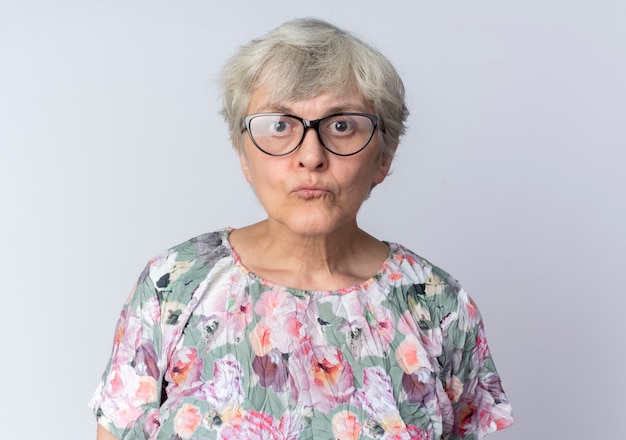 Shocked elderly woman in optical glasses stands isolated on white wall