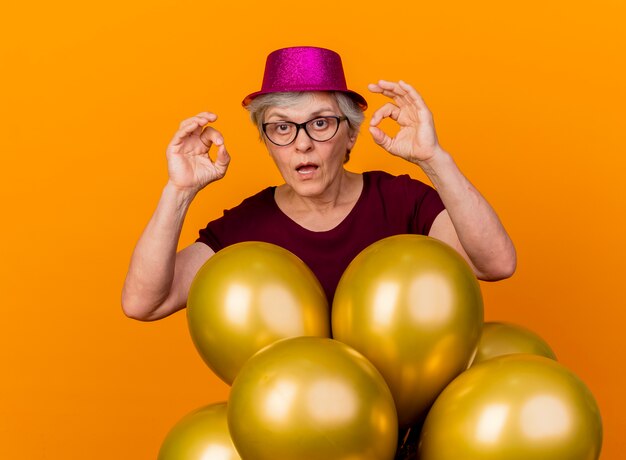 파티 모자를 쓰고 광학 안경에 충격을받은 노인 여성은 헬륨 풍선이 주황색 벽에 고립 된 두 손으로 확인 손 기호를 몸짓으로 의미합니다.