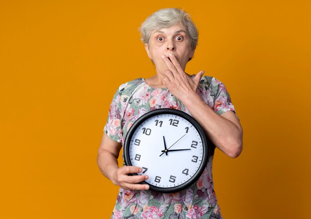 Shocked elderly woman puts hand on mouth holding clock isolated on orange wall