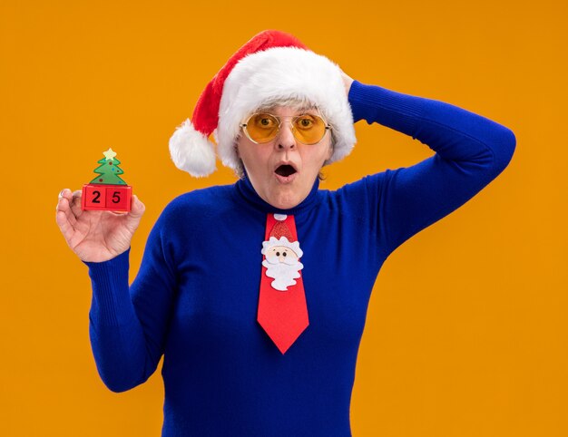 Shocked elderly woman in sun glasses with santa hat and santa tie holds christmas tree ornament and puts hand on head isolated on orange background with copy space