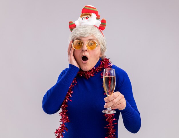 Shocked elderly woman in sun glasses with santa headband and garland around neck puts hand on face and holds glass of champagne 