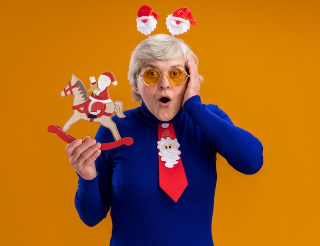 Free photo shocked elderly woman in sun glasses with santa headband and santa tie holding santa on rocking horse decoration and putting hand on face isolated on orange background with copy space