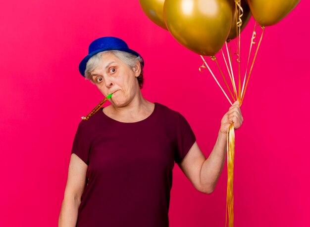 Shocked elderly woman wearing party hat holds helium balloons blowing whistle on pink