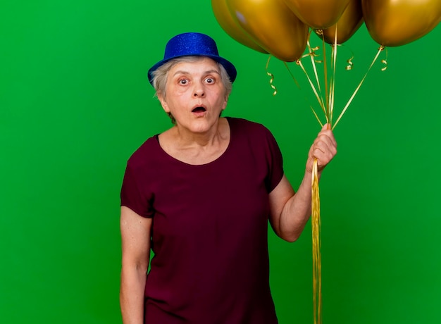 Foto gratuita la donna anziana scioccata che porta il cappello del partito tiene i palloni dell'elio sul verde