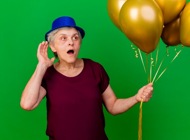 Shocked elderly woman wearing party hat holds helium balloons keeping hand behind ear looking at side on green