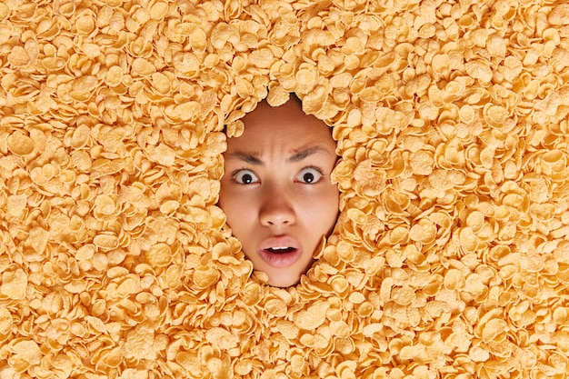 Shocked embarassed woman stares with indignant expression drowned in dry cornflakes has balanced nutrition and dietary ration