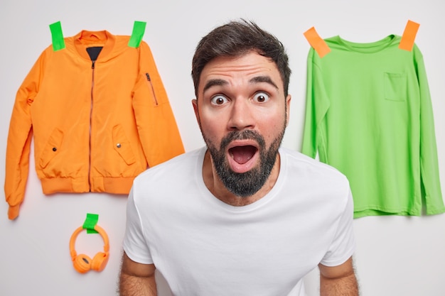 Shocked embarrassed bearded adult man stares at front keeps mouth wide opened from amazement cannot believe in shocking news poses indoor with clothes items plastered to wall in wall