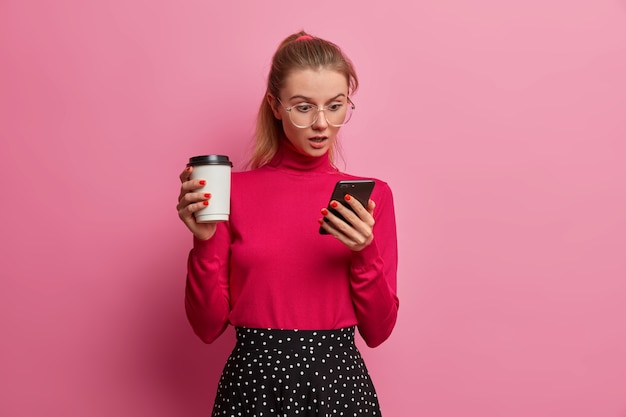 Shocked emotional girl stares at smartphone display, chats with friends online, wears big optical glasses, holds disposable cup of fresh drink, enjoys tasty coffee