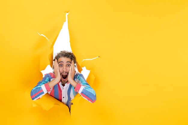 Shocked and emotional young guy posing for camera through a torn hole in yellow paper