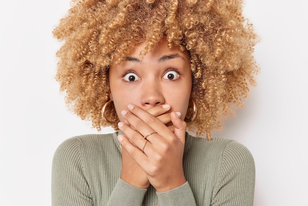 Shocked emotional young woman with curly hair covers mouth with hands tries to be speechless reacts on horrible scene wears casual turtleneck isolated over white background Unbelievable news