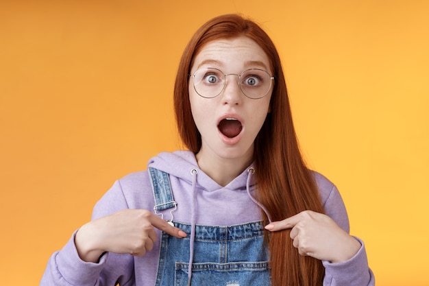 Shocked emotive surprised redhead european woman wearing glasses hoodie drop jaw gasping stunned pointing herself full disbelief cannot believe win being picked look impressed orange background