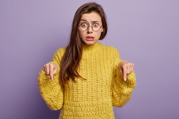 Shocked European lady looks with puzzlement, being speechless, points down on floor, has nervous expression, stares through spectacles, dressed in yellow jumper, isolated on purple wall