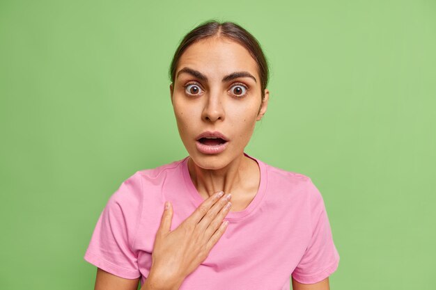 Shocked European woman holds breath keeps mouth opened cannot believe in shocking breathtaking news wears casual clothes isolated over green wall