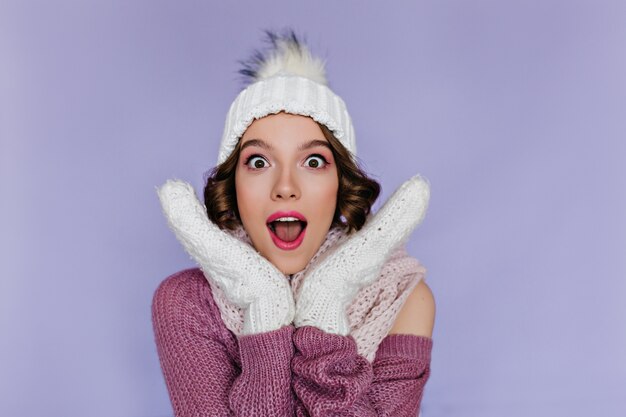 Shocked european woman wears soft white mittens posing  Indoor portrait of short-haired female model wears woolen hat and gloves.