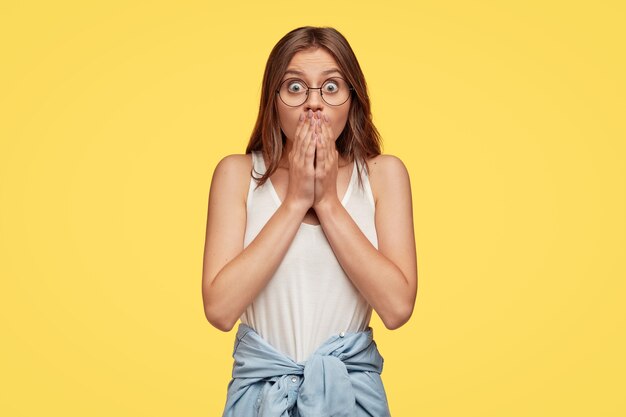 Shocked European woman with long dark hair, keeps hands on mouth, stares through round spectacles