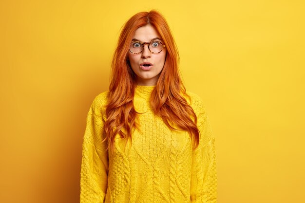 Shocked European woman with red natural hair keeps mouth opened has stunned expression holds breath dressed in knitted sweater.