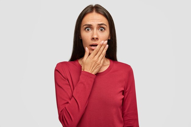 Shocked European young attractive female with dark hair, keeps hand near mouth, stares with bugged eyes as hears awful news from interlocutor, wears red sweater, isolated over white wall.