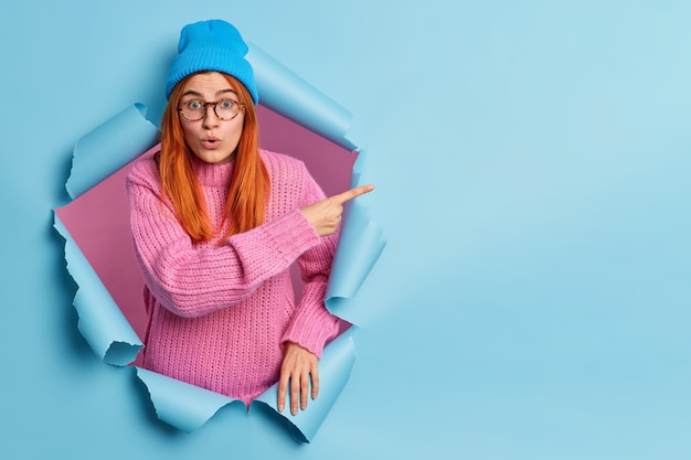 Free photo shocked excited redhead teenage girl peeps through hole in paper pointing at copy space