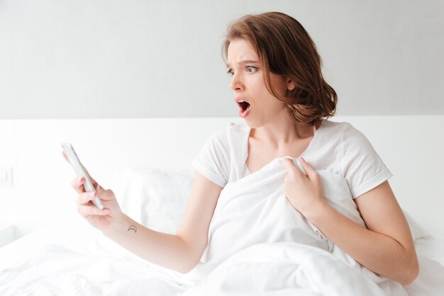 携帯電話でおしゃべりベッドでショックを受けて興奮している若い女性。