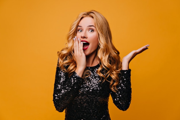 Shocked fair-haired girl posing on yellow wall with mouth open.  surprised glamorous lady.