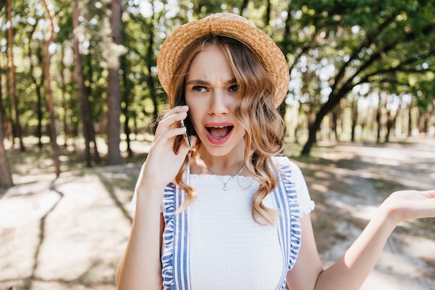 Shocked fair-haired girl standing in park and talking on phone. Outdoor shot of beautiful young woman in hat expressing amazement during conversation.