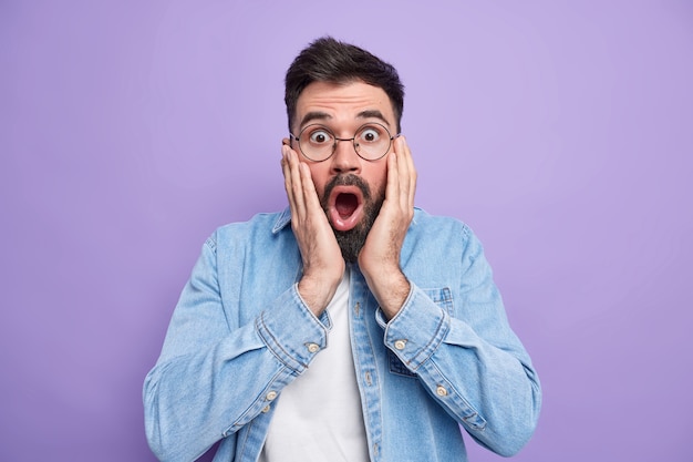 Free photo shocked fascinated man keeps hands on face wide opened mouth checks out something terrible stares through spectacles wears denim shirt