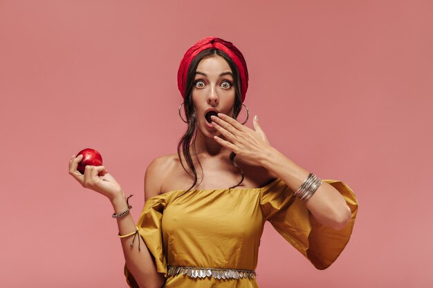 Shocked fashionable woman with cool accessories and yellow dress looking into camera and holding red apple on pink wall