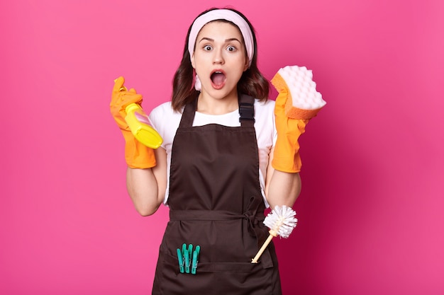 Shocked female housekeeper holds sponge and detergent spray in hands, having much work to do. Attractive woman with surprised and excited look wearing apron and protective gloves. Copy space.