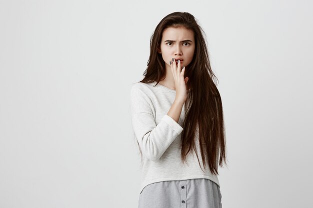 Shocked female in loose shirt with surprised expression, finds out unexpected news, can t believe in words she is told, says is it really so Emotional woman closes mouth with hand in excitement