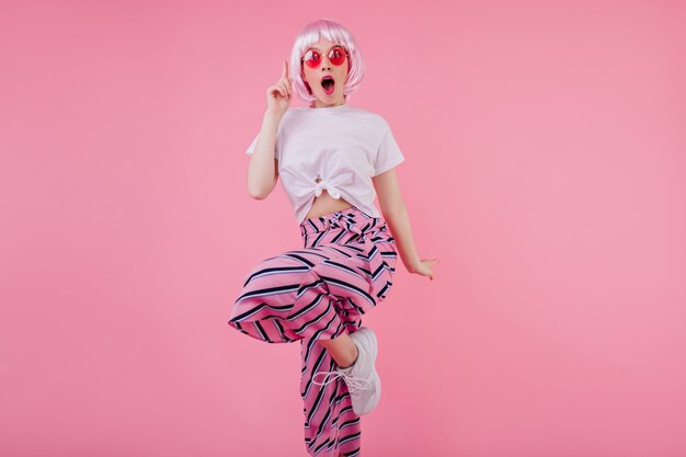 Шокированная женская модель в элегантном розовом перуке танцует в модной одежде. Крытая изысканная девушка в парике, выражающая удивленные эмоции