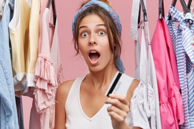 Shocked female in white T-shirt, standing near sale clothes rack, holding credit card, being surprised that she has no money on her accountant, wanting to refresh her wardrobe. Buying outfit