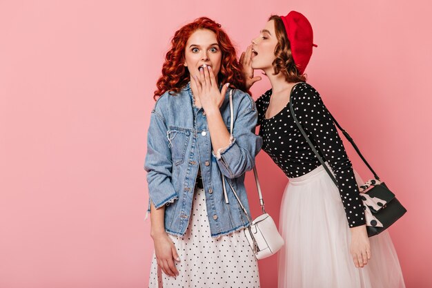 Shocked ginger girl listening friend. Two women talking on pink background.